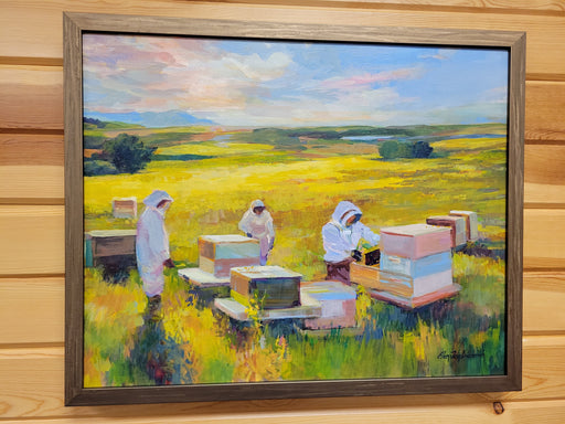 A framed landscape oil painting depicting beekeepers in white suits working with beehives in a yellow field under a blue and pink sky, mounted on a light wood paneled wall.