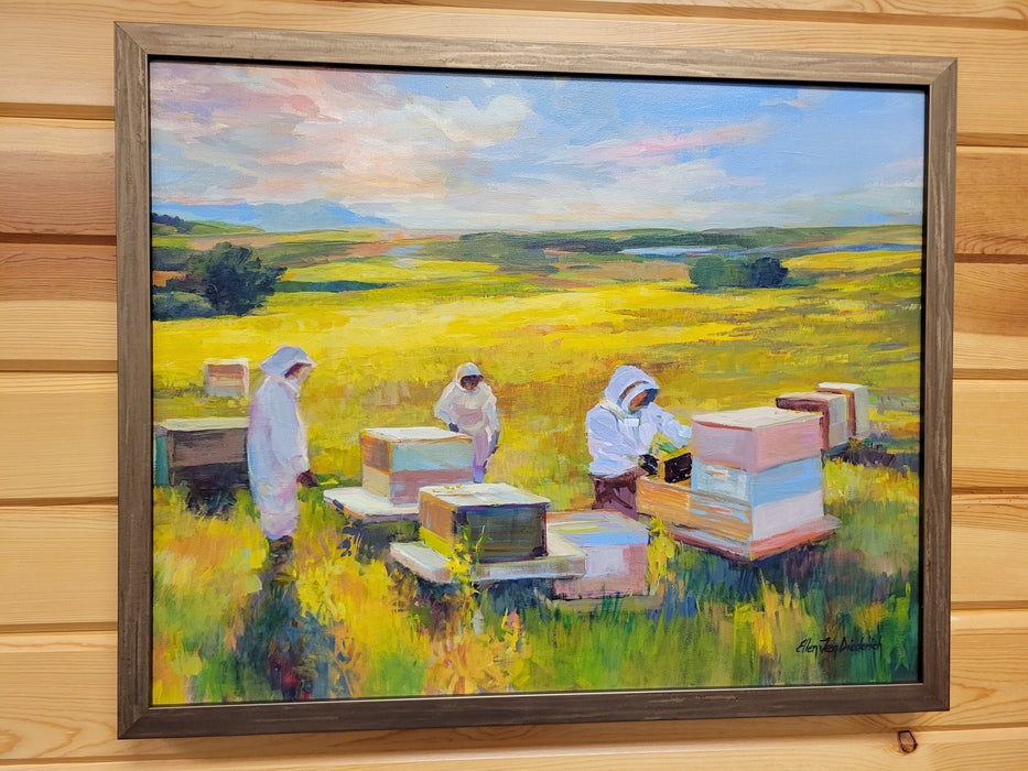 A framed landscape oil painting depicting beekeepers in white suits working with beehives in a yellow field under a blue and pink sky, mounted on a light wood paneled wall.
