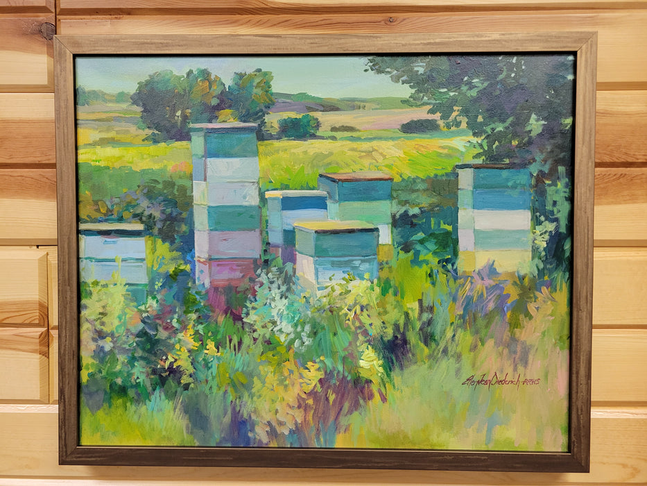 A framed landscape oil painting depicting beehives in a field, mounted on a light wood paneled wall. The painting features green, blue, and yellow tones with a dark brown wooden frame.